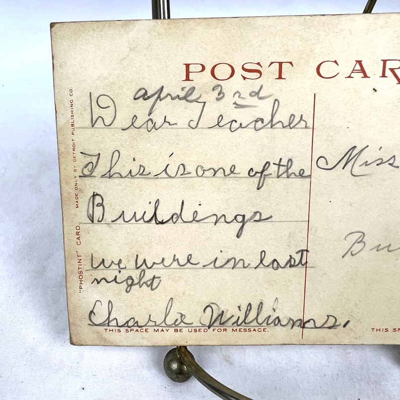 May include: Aged postcard with a handwritten message addressed to "Dear Teacher" dated 3rd April. The message mentions "Buildings" and is signed "Charla Williams." The postcard is held by a decorative metal stand.
