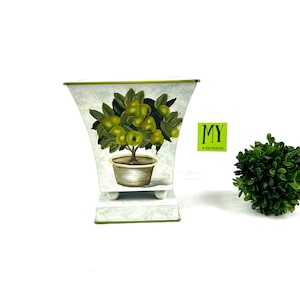 以下が含まれることがあります： 緑色の果物と葉が付いた鉢植えの果樹の塗装デザインが施された装飾的な金属製のプランター。 プランターは白で緑色のトリムがあり、白いベースの上にあります。 テキスト「MY 90 YEAR COLLECTION」は緑色のステッカーにあります。