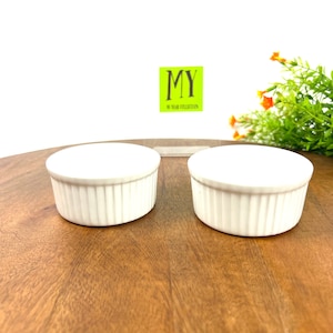 May include: Two white ceramic ramekins with fluted edges, sitting on a wooden surface. The ramekins are empty and ready to be used for baking or serving.