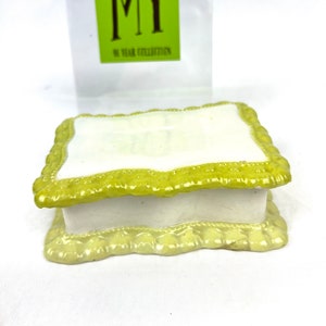 May include: A small, white ceramic box with a yellow, decorative trim. The box has a lid and is decorated with a scalloped edge.