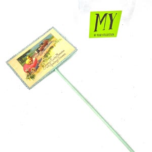 May include: A vintage Easter greeting on a stick. The rectangular card features a colorful illustration of a chick in a carriage and the text "Joyous Easter Memories with Greetings Simeere." The stick is light green.