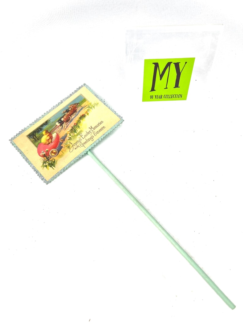 May include: A vintage Easter greeting on a stick. The rectangular card features a colorful illustration of a chick in a carriage and the text "Joyous Easter Memories with Greetings Simeere." The stick is light green.
