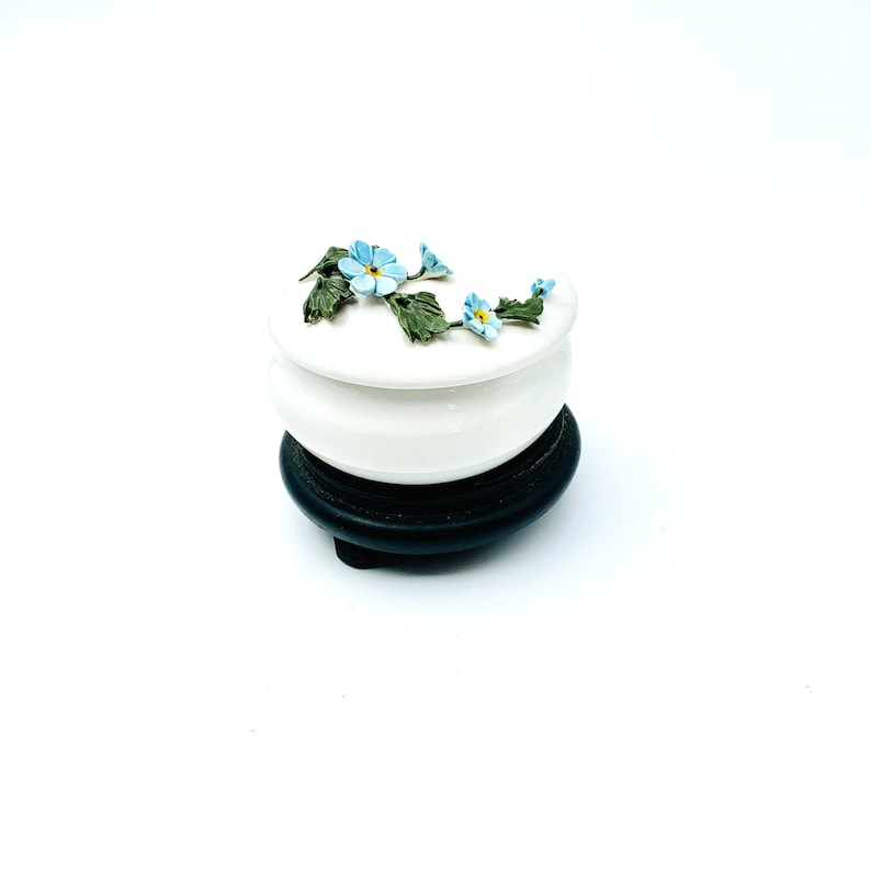 May include: A white ceramic trinket box with a floral design featuring blue forget-me-not flowers and green leaves. The box sits on a black wooden base.