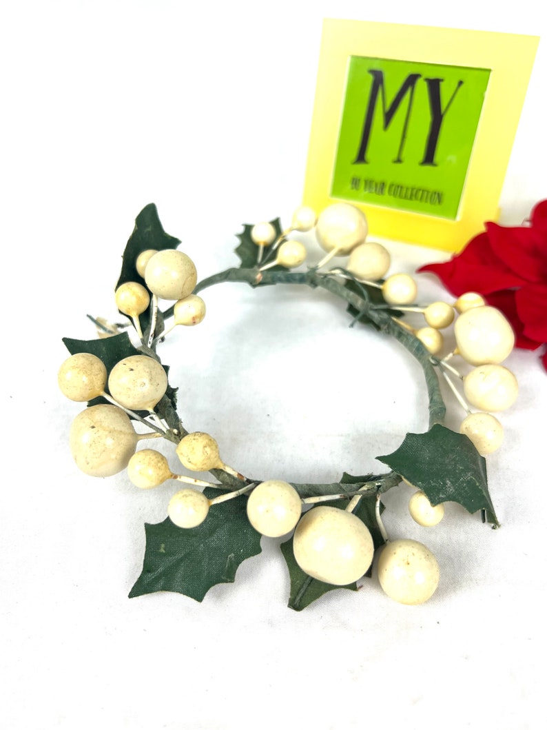 May include: A white and green artificial holly wreath with white berries. The wreath is made of wire and has a simple design.