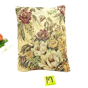 May include: A decorative throw pillow with a floral pattern in shades of brown, yellow, and green. The pillow has a cream-colored background and features a variety of flowers, including roses, daisies, and tulips.