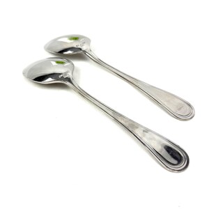 May include: Two shiny, silver spoons with oval bowls and long, slender handles. The spoons are set against a white background, with the light reflecting off the polished surfaces. The spoons appear to be part of a flatware set.