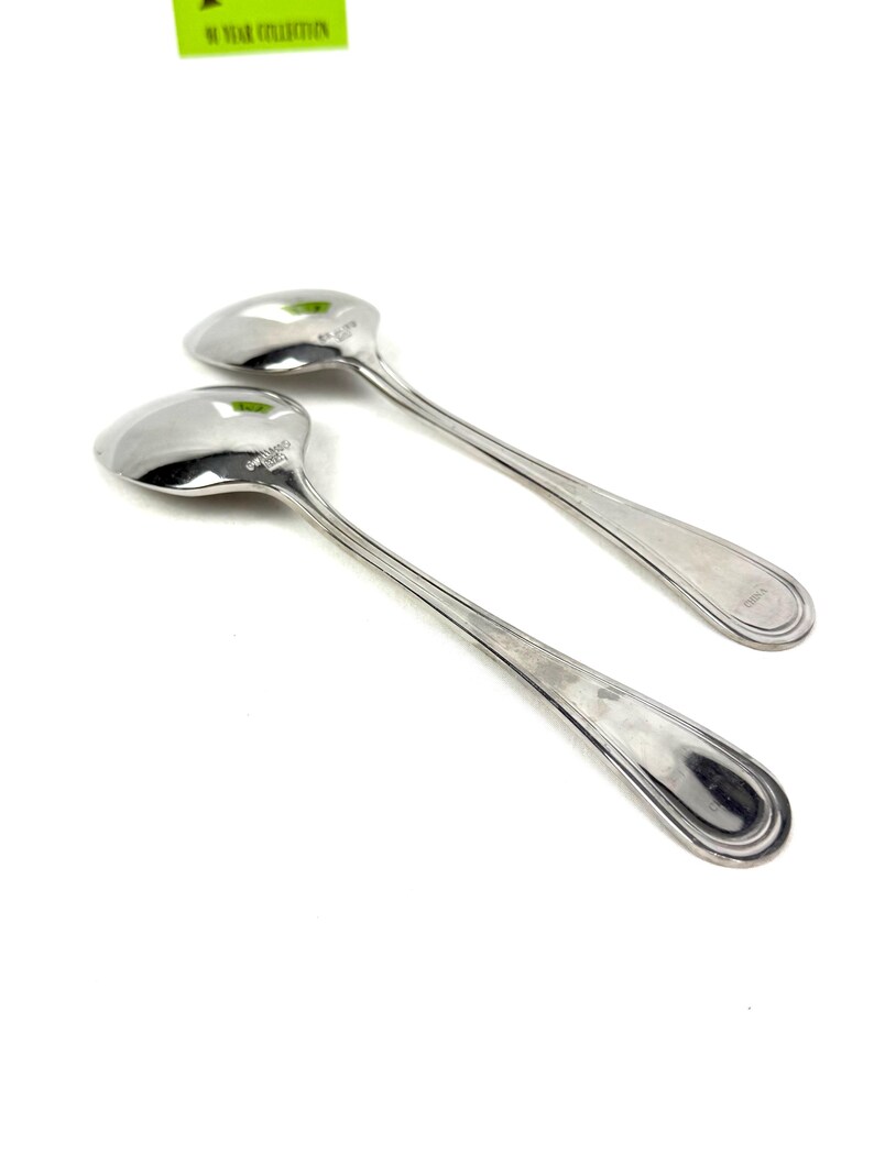 May include: Two shiny, silver spoons with oval bowls and long, slender handles. The spoons are set against a white background, with the light reflecting off the polished surfaces. The spoons appear to be part of a flatware set.