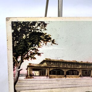 May include: A vintage postcard showing a building with arched openings and a balcony. A large tree with dark green leaves is on the left. The postcard has a faded look, with a light grey sky in the background.