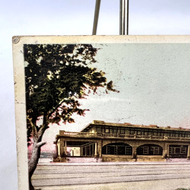 May include: A vintage postcard showing a building with arched openings and a balcony. A large tree with dark green leaves is on the left. The postcard has a faded look, with a light grey sky in the background.