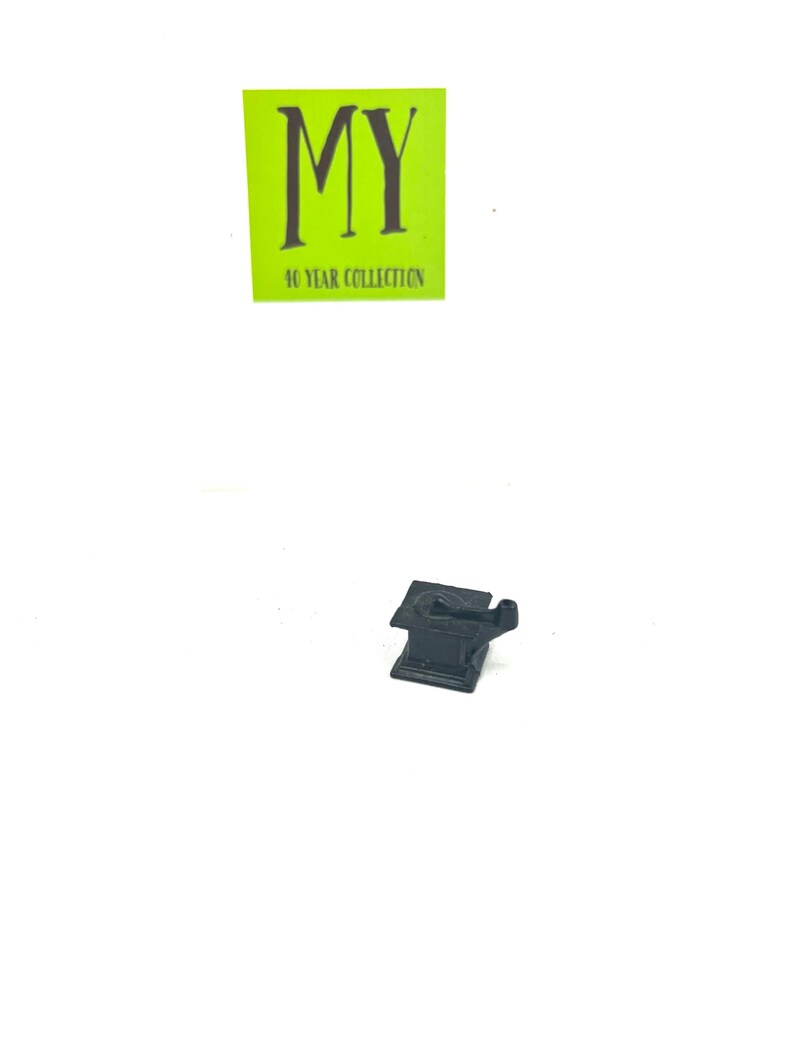 May include: A black metal miniature anvil with a small hammer on top. The anvil is sitting on a white surface.