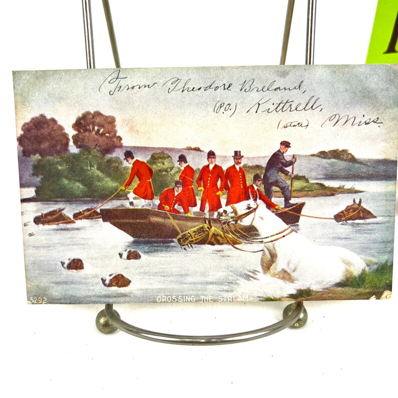 K&ouml;nnte beinhalten: Eine antike Postkarte, die eine Gruppe von M&auml;nnern in roten M&auml;nteln und Zylindern zeigt, die Pferde durch einen Fluss f&uuml;hren. Die Postkarte tr&auml;gt den Titel "Crossing the Stream" und ist signiert mit "From Theodore Breland, (P.O.) Kittrell, (state) Miss."