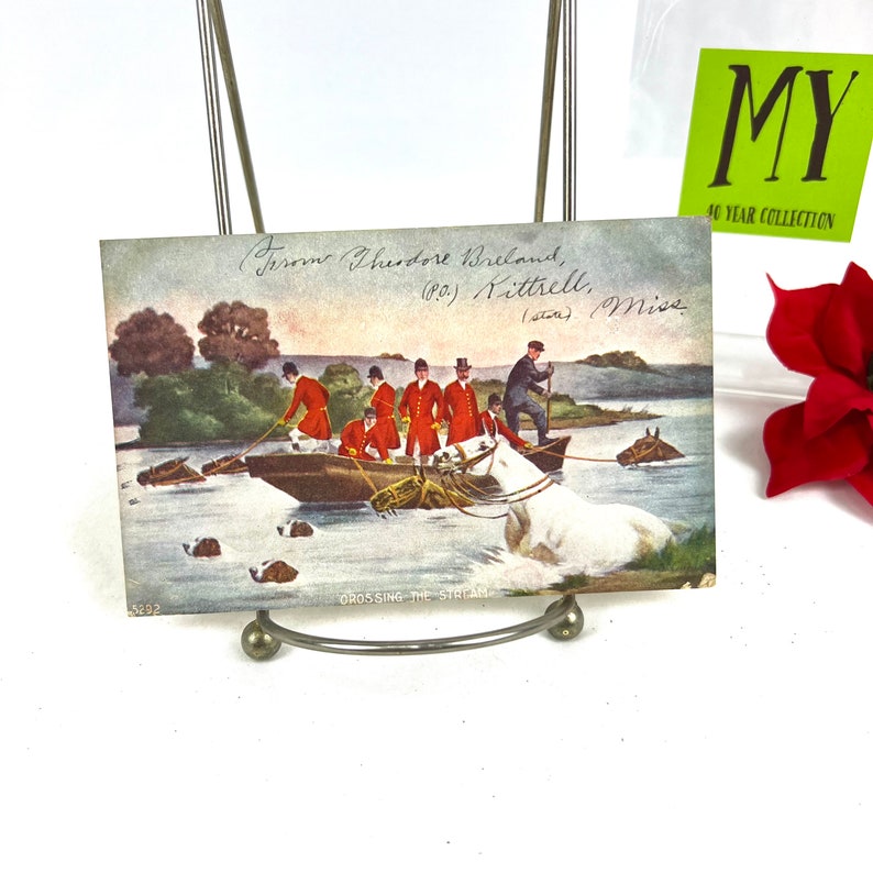 K&ouml;nnte beinhalten: Vintage-Postkarte, die eine Gruppe von M&auml;nnern in roten M&auml;nteln und Zylindern zeigt, die in einem Boot einen Bach &uuml;berqueren, w&auml;hrend Hunde nebenher schwimmen. Die Postkarte tr&auml;gt den Titel "Crossing the Stream" und eine handschriftliche Inschrift auf der R&uuml;ckseite.