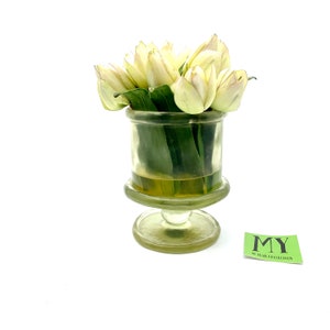May include: A clear glass vase with a pedestal base holds a bouquet of white tulips. The vase has a textured surface and the tulips have green leaves.