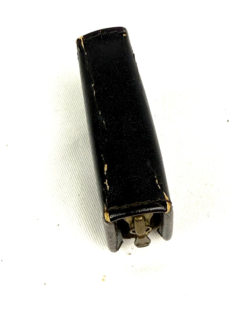May include: A black leather case with a brass zipper closure. The rectangular case is slightly worn, with visible stitching and wear on the edges. The zipper is a vintage style, adding to the overall aesthetic.
