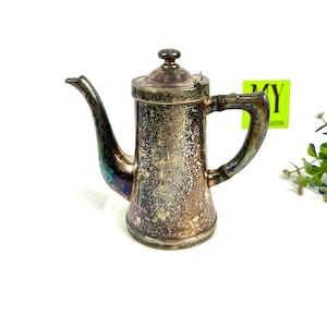 Op de afbeelding: Antieke zilverkleurige koffiepot met een gebogen tuit en een handvat. De pot heeft een deksel en een aangetaste afwerking, waardoor hij een vintage uitstraling heeft. De pot is waarschijnlijk gemaakt van metaal en is ontworpen voor het zetten van koffie.