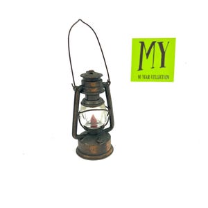 May include: A miniature antique-style lantern with a clear glass globe and a bronze-colored metal frame. The lantern has a handle for carrying and a small red light inside. A green sticker with the text "MY 10 YEAR COLLECTION" is in the background.