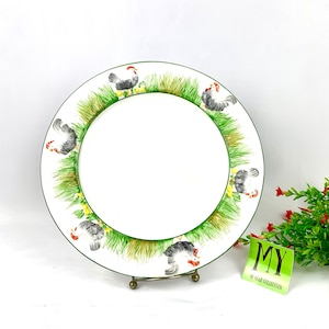 May include: A white ceramic dinner plate with a green border and a design of black and white chickens in a grassy field. The plate is on a metal stand.