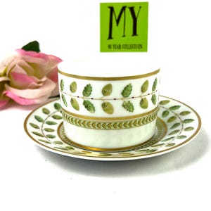 May include: A white porcelain teacup and saucer set with a green and gold leaf design. The saucer has a gold rim and the cup has a gold band around the top. The cup and saucer are decorated with a repeating pattern of green leaves and gold accents.