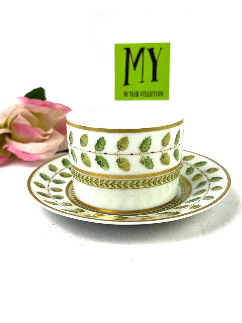 May include: A white porcelain teacup and saucer set with a green and gold leaf design. The saucer has a gold rim and the cup has a gold band around the top. The cup and saucer are decorated with a repeating pattern of green leaves and gold accents.