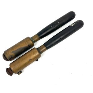 May include: Two antique metal tools with black handles and brass-colored bodies. The tools have a cylindrical shape with a rounded end on the handle and a smaller, rounded end on the opposite side. The tools are lying on a white surface.