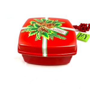 May include: Red square tin box with a Christmas design featuring holly, pine branches, pine cones, and a white ribbon with the text "Season's Greetings".