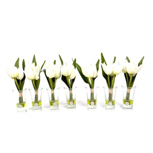 Peut inclure: Sept bouquets de tulipes blanches dans des verres &agrave; shot transparents avec des accents verts et argent&eacute;s. Chaque tulipe a des feuilles vertes et est attach&eacute;e avec de la ficelle. Les verres &agrave; shot sont dispos&eacute;s en rang&eacute;e sur fond blanc.