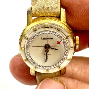 May include: A vintage Lucerne Swiss watch with a gold-tone case and a cream-colored face. The watch face features a ring of small crystals, black hands, and the text "Lucerne" and "Swiss". The watch has a light brown leather band.