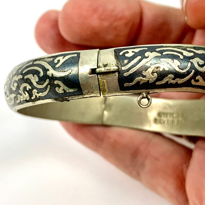 May include: A silver bangle bracelet with a black enamel design and a filigree pattern. The bracelet is hinged and has a small charm hanging from it. The inscription "STERLING SILVER" is visible on the bracelet.