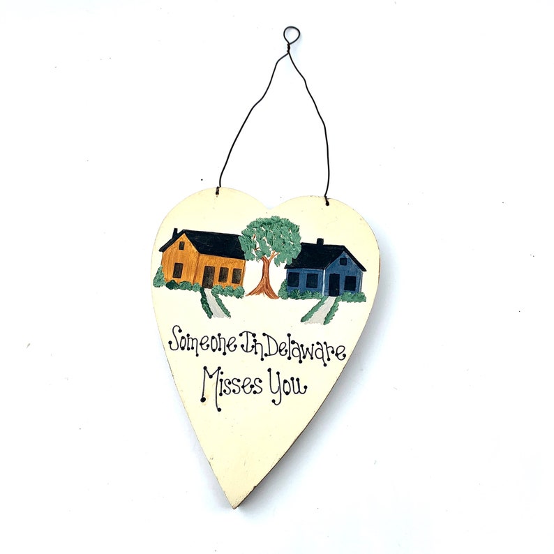May include: A white heart-shaped wooden sign with a painted scene of two houses and a tree. The text "Someone In Delaware Misses You" is written in black cursive.
