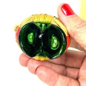 May include: Green glass bottle stopper with a woven straw and red trim. The stopper is shaped like a circle with a small opening in the center.
