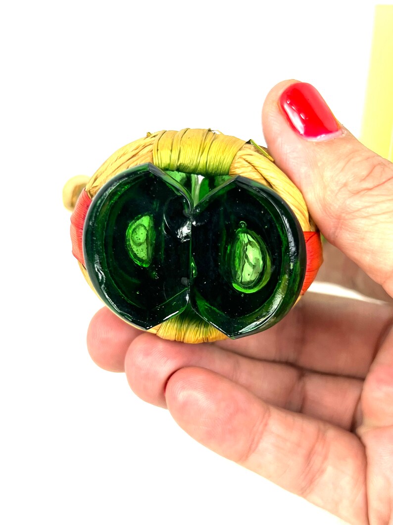May include: Green glass bottle stopper with a woven straw and red trim. The stopper is shaped like a circle with a small opening in the center.