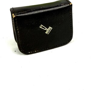 May include: A small, black leather case with a gold-colored emblem on the front. The case is rectangular with rounded edges and appears to be closed. The emblem is a stylized design, possibly a logo or symbol.