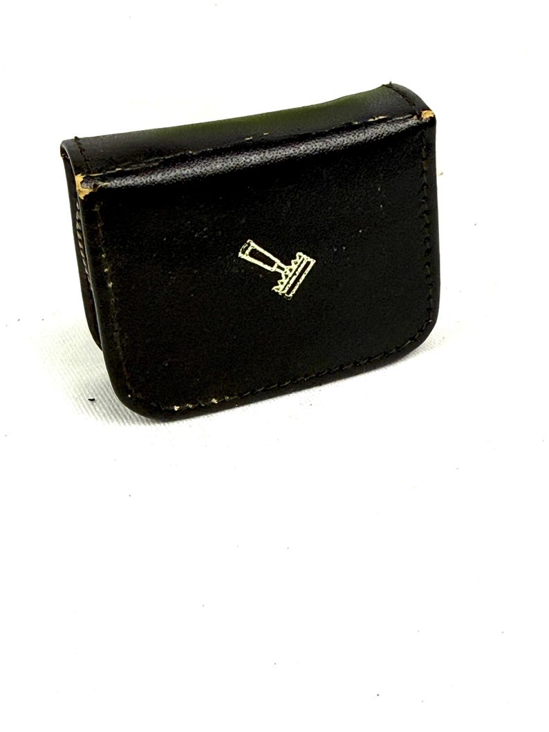 May include: A small, black leather case with a gold-colored emblem on the front. The case is rectangular with rounded edges and appears to be closed. The emblem is a stylized design, possibly a logo or symbol.
