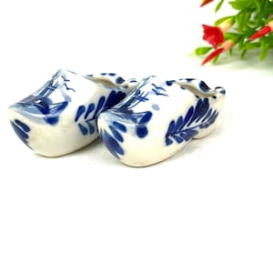 May include: A pair of miniature ceramic clogs, painted white with blue floral and geometric patterns. The clogs are displayed on a white surface. The image includes a green and red floral arrangement and a green label that reads "40 YEAR COLLECTION".