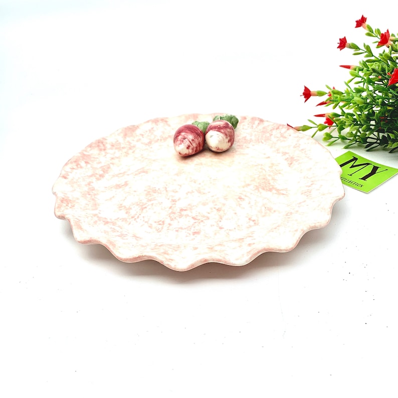 May include: A pink ceramic plate with a scalloped edge. The plate has a marbled design and is decorated with two small, red and green ceramic strawberries.