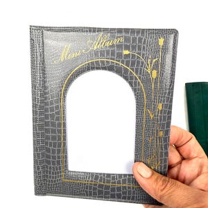 Peut inclure: Un album photo gris avec un motif textur&eacute; en forme de peau de crocodile. L'album pr&eacute;sente un cadre dor&eacute; en forme d'arche autour de l'ouverture de la photo et des accents floraux dor&eacute;s. Les mots "Mem-Album" sont en lettres dor&eacute;es en haut.