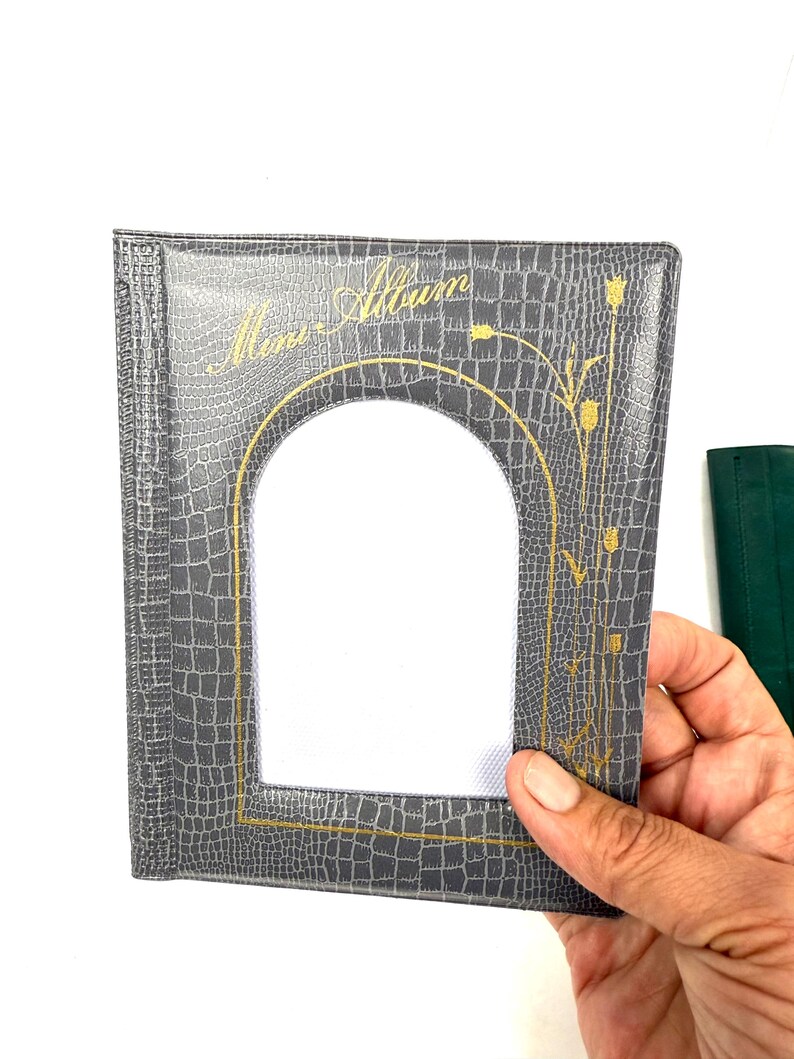 Peut inclure: Un album photo gris avec un motif textur&eacute; en forme de peau de crocodile. L'album pr&eacute;sente un cadre dor&eacute; en forme d'arche autour de l'ouverture de la photo et des accents floraux dor&eacute;s. Les mots "Mem-Album" sont en lettres dor&eacute;es en haut.