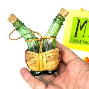 May include: Two small green glass bottles with cork stoppers are tied together with twine and wrapped in woven straw. The bottles are in a woven basket with red accents.