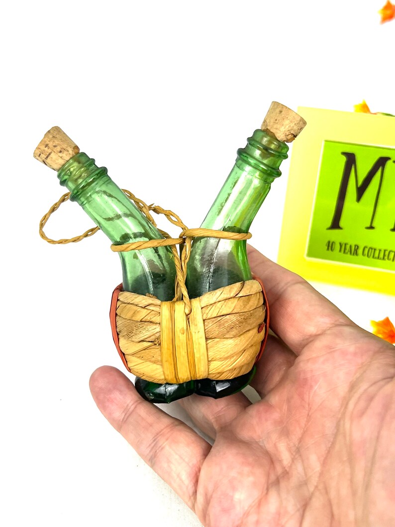 May include: Two small green glass bottles with cork stoppers are tied together with twine and wrapped in woven straw. The bottles are in a woven basket with red accents.