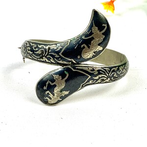 May include: A silver bangle bracelet with a black enamel finish. The bracelet features two silver figures of dancers in a traditional Thai style. The bracelet is decorated with intricate silver filigree.