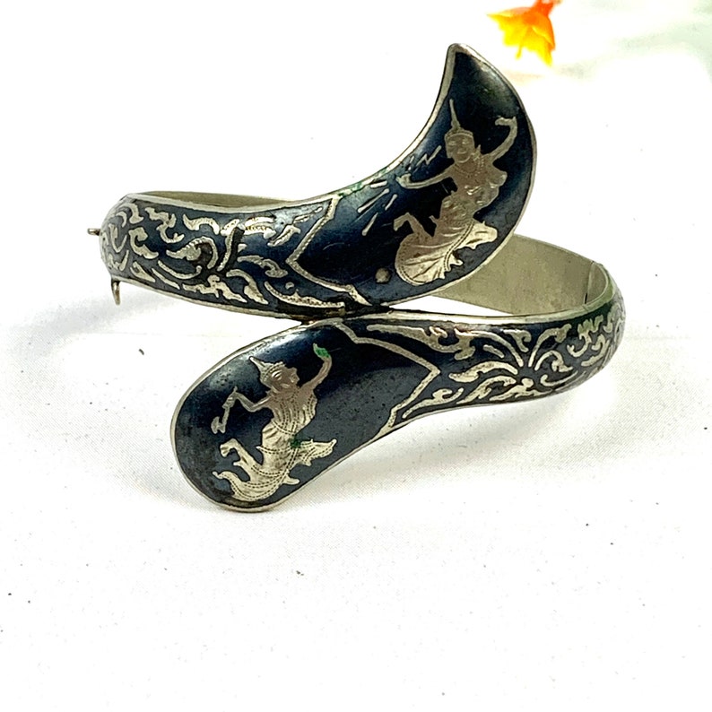 May include: A silver bangle bracelet with a black enamel finish. The bracelet features two silver figures of dancers in a traditional Thai style. The bracelet is decorated with intricate silver filigree.
