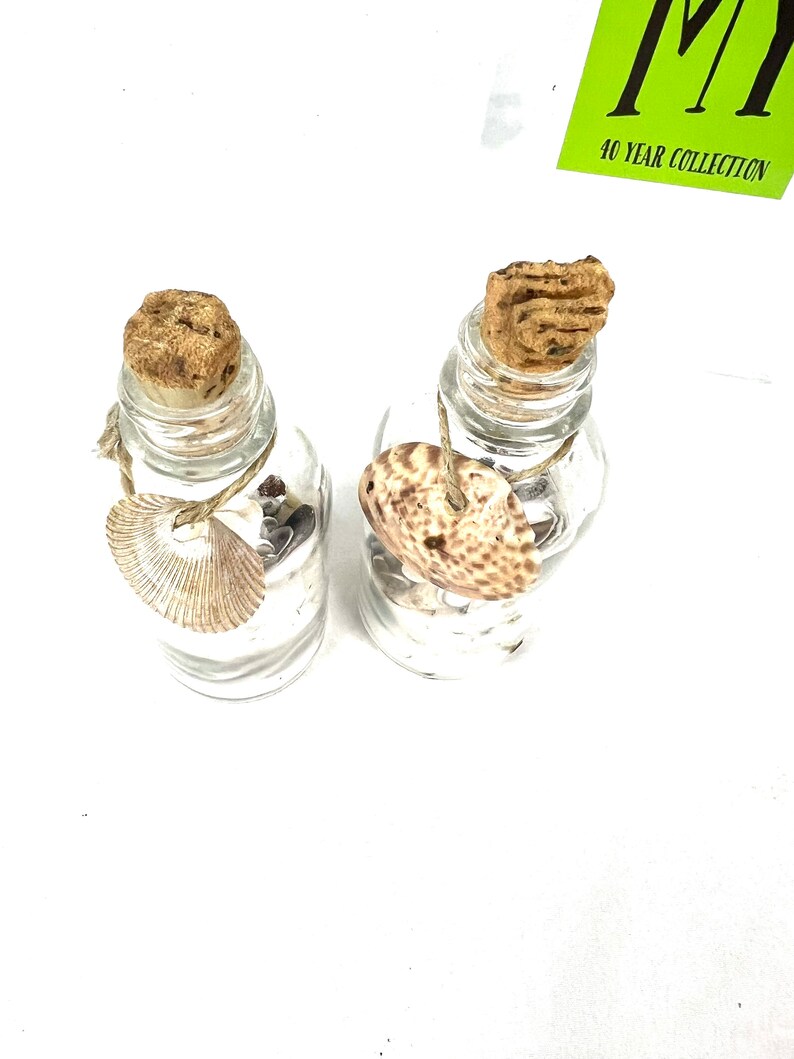 May include: Two small glass bottles with cork stoppers, each filled with sand and a seashell. The bottles are tied with twine and have a rustic, beachy aesthetic.