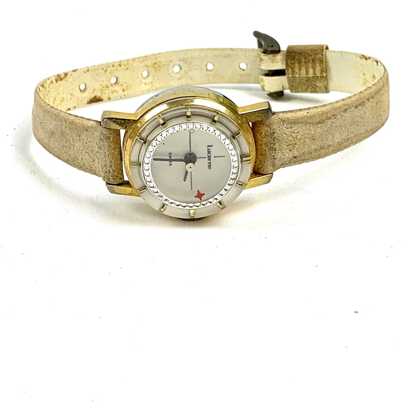 May include: Vintage Lucerne Swiss watch with a gold-tone case and a light-colored leather band. The watch face is silver with a diamond-like bezel and a red star emblem. The band is approximately 0.5 inches wide.