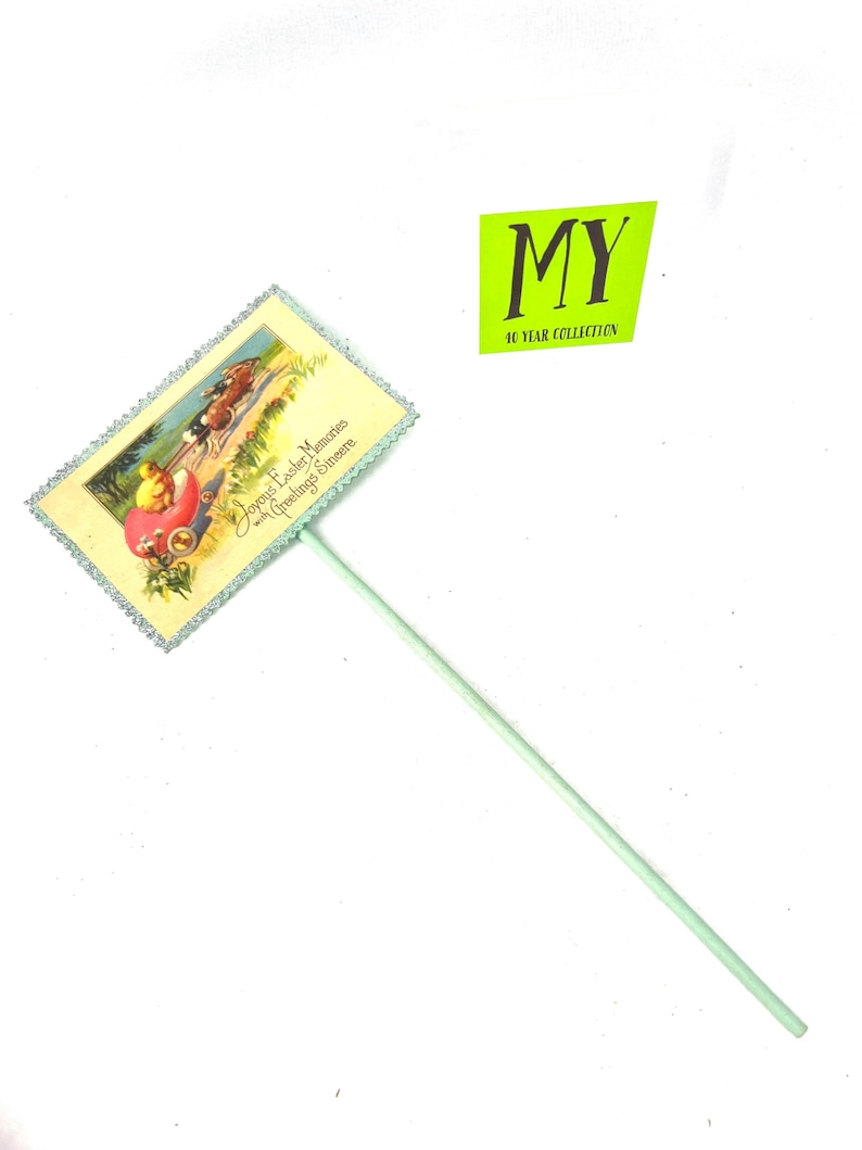 May include: A vintage Easter-themed cake topper featuring a pastel illustration of a chick in a carriage pulled by horses. The topper has a light green stick and a decorative border. The text on the topper reads "Joyous Easter Memories with Greetings Sincere."