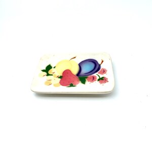 May include: A white ceramic dish with a painted design of fruit, including a yellow pear, a purple plum, a red strawberry, and pink flowers.