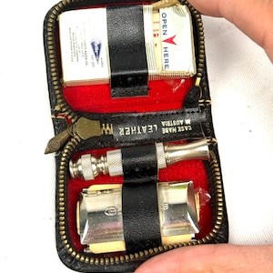 May include: A black leather travel kit with a gold zipper, red interior, and black straps. The kit contains a razor, a shaving brush, and a box of matches. The text "CASE MADE IN AUSTRIA LEATHER" is visible.