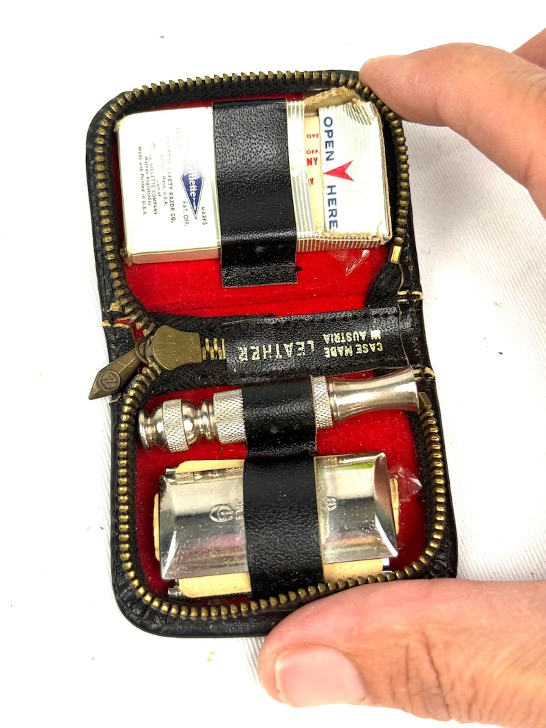 May include: A black leather travel kit with a gold zipper, red interior, and black straps. The kit contains a razor, a shaving brush, and a box of matches. The text "CASE MADE IN AUSTRIA LEATHER" is visible.