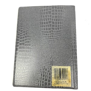 Peut inclure: Un cahier rectangulaire gris avec un motif textur&eacute; en forme de peau de crocodile. Une &eacute;tiquette avec un code-barres et du texte est appos&eacute;e en bas &agrave; droite. Le cahier est ferm&eacute; et semble neuf.