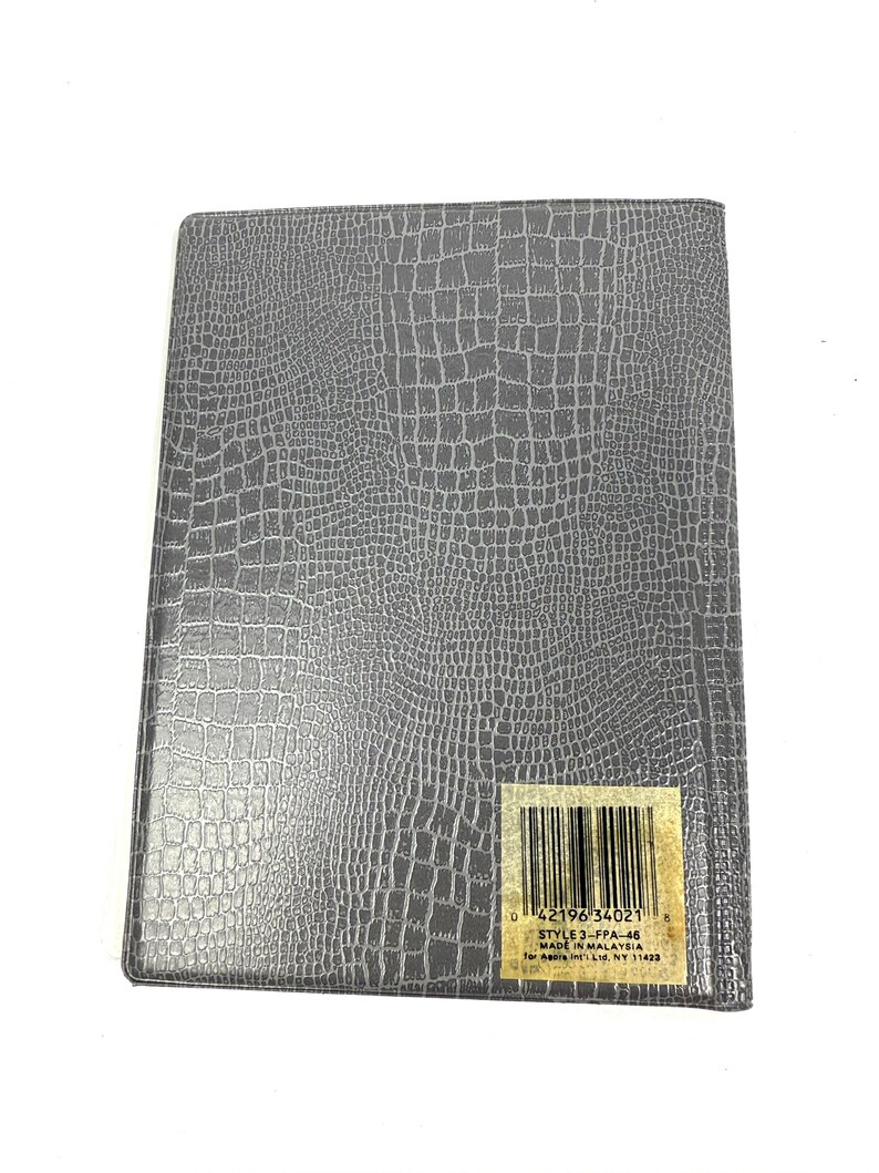Peut inclure: Un cahier rectangulaire gris avec un motif textur&eacute; en forme de peau de crocodile. Une &eacute;tiquette avec un code-barres et du texte est appos&eacute;e en bas &agrave; droite. Le cahier est ferm&eacute; et semble neuf.