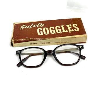 Pode incluir: &Oacute;culos de seguran&ccedil;a vintage com uma caixa castanha. A caixa diz "Safety Goggles" em letras brancas. Os &oacute;culos s&atilde;o pretos com lentes transparentes e um desenho de cruz nos cantos. O texto "PROTECT YOUR EYES... wear goggles!" est&aacute; na caixa.